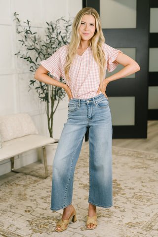 A woman modeling a pink and white gingham short-sleeve blouse with ruffled sleeves, paired with light-wash wide-leg blue jeans and tan heeled sandals, standing in a bright, modern room.
