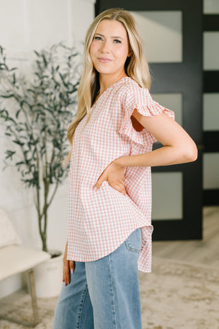 A woman modeling a pink and white gingham short-sleeve top with ruffled sleeves, paired with light blue jeans in a bright indoor setting.