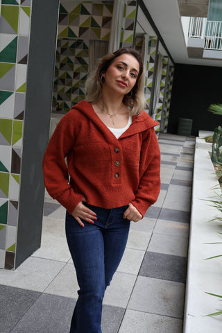 Woman wearing a red sweater and blue jeans standing in a modern indoor setting with geometric wall patterns.