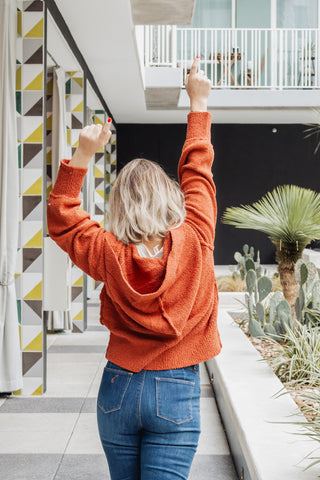 Person in an orange sweater and blue jeans standing with arms raised in a modern indoor setting with plants.