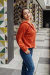 Woman wearing an orange sweater and blue jeans standing in a modern indoor setting with geometric wall patterns.