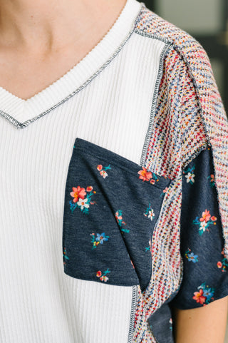Close-up of a person wearing a white ribbed V-neck top with a large dark navy floral chest pocket and a multicolor knit cardigan draped over one shoulder.