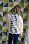 Person wearing a white cardigan over a black and white patterned shirt against a geometric tile wall.