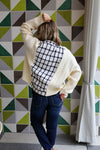 Person wearing a white sweater with a black and white patterned back, standing against a geometric tile wall.