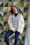 Woman wearing a cream cardigan over a patterned shirt against a geometric tile wall.