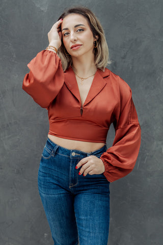 Woman wearing a rust-colored blouse and blue jeans against a gray background