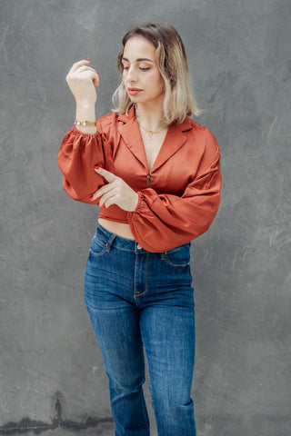 Woman wearing a rust-colored blouse and blue jeans against a gray background