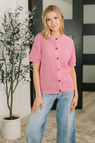 A woman wearing a pink short-sleeve knit cardigan with brown buttons and light blue jeans, standing indoors in front of a dark paneled door and a potted plant.