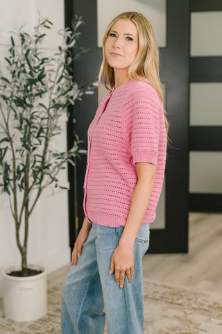 Woman wearing a pink sweater and blue jeans standing indoors next to a plant.