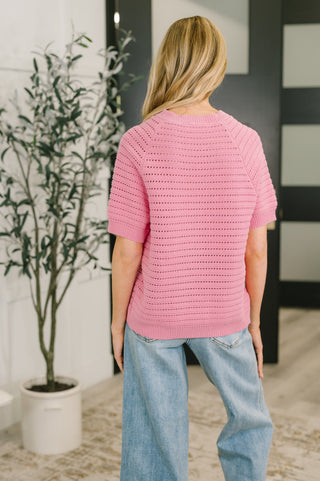 Person wearing a pink knitted sweater and blue jeans standing in a room with a plant and wall.