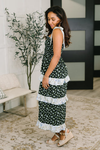 A woman wearing a sleeveless black midi dress with a white floral print and tiered ruffle details, standing in a modern indoor setting with a bench, potted plant, and patterned rug.