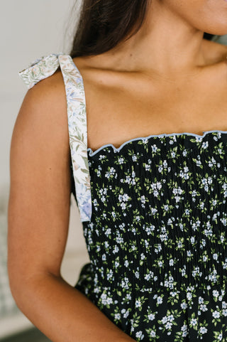 Close-up of a person wearing a smocked black floral dress with white and green flowers, featuring a light blue scalloped edge and a wide floral shoulder strap with a bow.