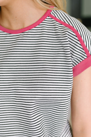 Close-up of a short-sleeve ringer T-shirt with thin black horizontal stripes on a white base and bright pink trim along the crew neck and sleeve cuffs, worn by a model.