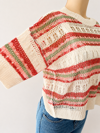 A close-up of a cream-colored open-knit sweater with horizontal stripes in red and green, styled on a mannequin with blue jeans against a plain light background.