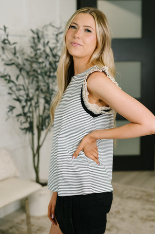 A woman modeling a sleeveless top with thin black and white horizontal stripes, featuring textured ruffle trim along the shoulder and armholes, paired with black shorts in an indoor setting.