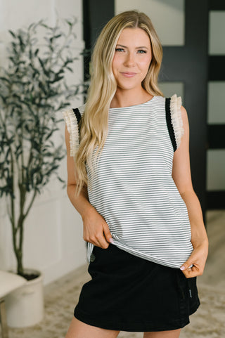 A woman modeling a sleeveless top with thin horizontal navy and white stripes, featuring textured cream ruffle trim along the shoulders and black trim along the armholes, paired with a black skirt.