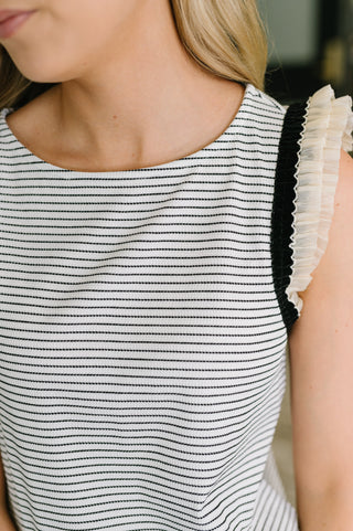 Close-up of a woman wearing a sleeveless top with thin horizontal black stripes on a white base and a ruffled off-white shoulder detail.