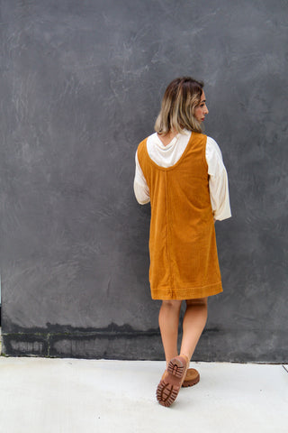 Woman wearing a mustard yellow pinafore dress against a gray wall.