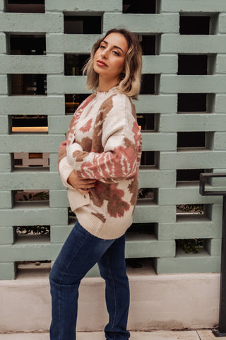 Woman wearing a tie-dye sweater and jeans standing against a textured wall.