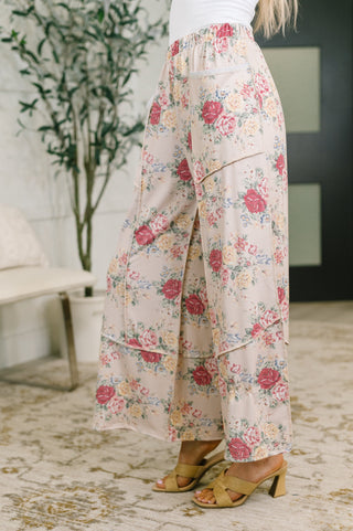 A person wearing wide-leg floral pants with a pink, red, and yellow rose pattern on a light beige background, paired with tan heeled sandals in an indoor setting.