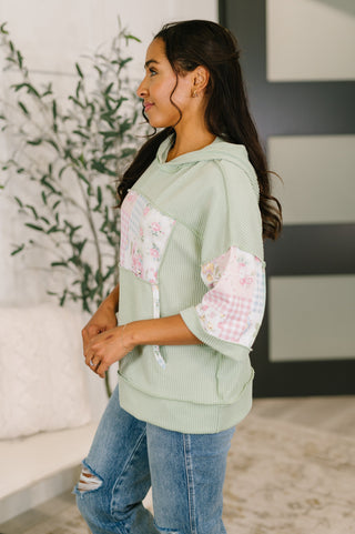 Woman wearing a light green hoodie with floral patch details in a room with a plant and abstract wall art.