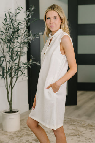 Woman wearing a white sleeveless dress standing indoors with a plant in the background