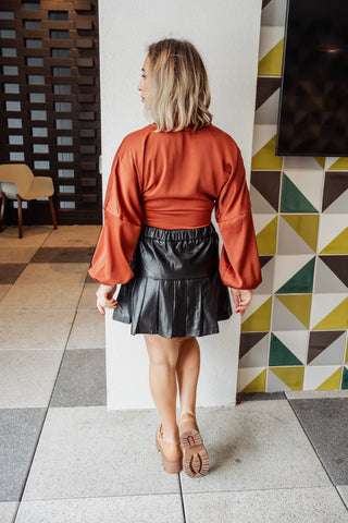 Woman wearing a rust-colored blouse and black leather skirt in a room with geometric-patterned wall.