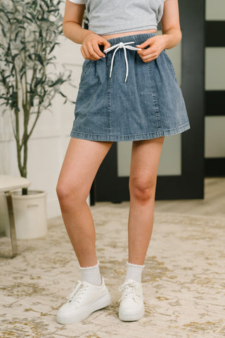 A person wearing a light blue denim mini skirt with a white drawstring waist, paired with a gray crop top, white socks, and white sneakers in an indoor setting.