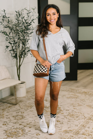A woman stands indoors wearing a light gray oversized V-neck T-shirt, denim shorts, white sneakers, and a black-and-white checkered crossbody bag with brown trim.