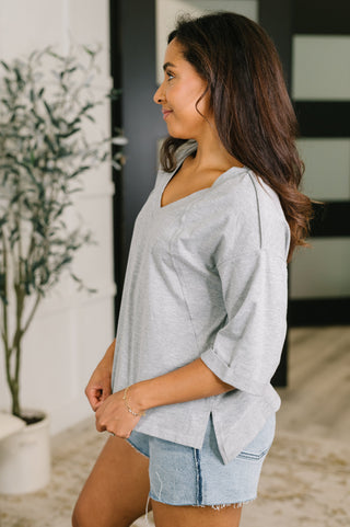 A woman wearing a light gray heathered V-neck top with three-quarter sleeves and light-wash denim shorts, standing in a modern indoor setting.