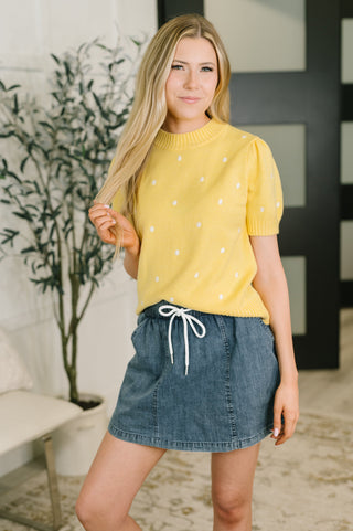 A woman modeling a yellow short-sleeve knit top with small white polka dots, paired with a denim mini skirt, in an indoor setting with a potted plant and modern wall decor.