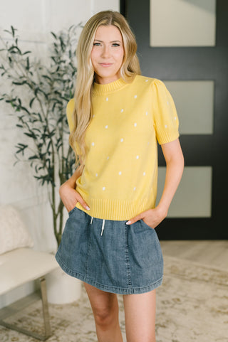 A woman modeling a bright yellow short-sleeve knit sweater with small white embroidered dots, paired with a denim skirt in an indoor setting.
