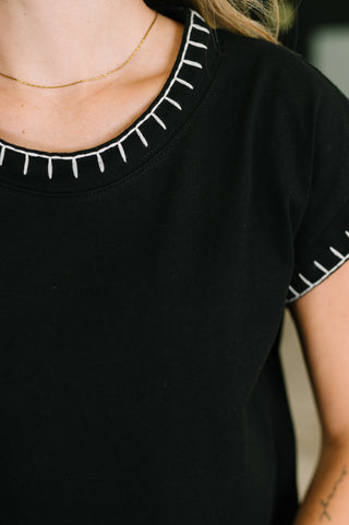 Close-up of a person wearing a black short-sleeve top with white embroidered or printed stitched details around the neckline and sleeve.