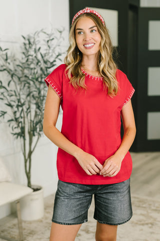 A woman wearing a bright red short-sleeve top with white contrast stitching along the neckline and sleeve hems, paired with gray denim shorts and a red-and-white patterned headband, standing in a modern indoor setting.