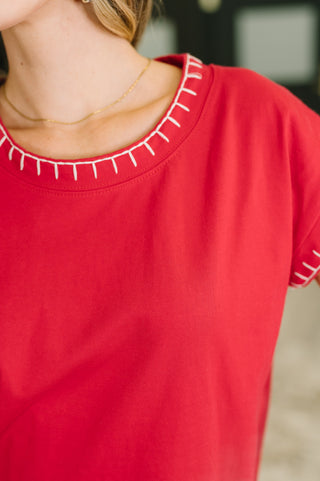 A close-up of a person wearing a bright red short-sleeve top with white decorative stitching around the crew neckline and sleeve cuffs.