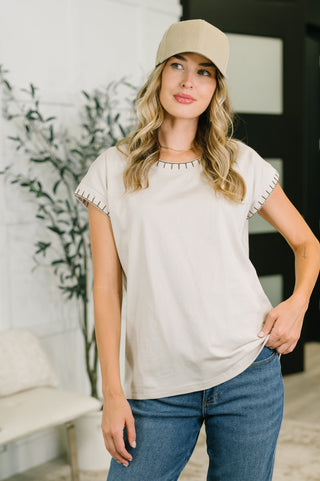 A woman wearing a light beige short-sleeve t-shirt with black scalloped trim on the sleeves, paired with blue jeans and a beige baseball cap, standing in a brightly lit indoor setting.