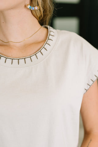 Close-up of a person wearing a light beige or off-white short-sleeve top with decorative dark stitched details around the neckline and sleeve edges.
