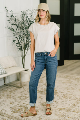 A woman stands indoors wearing a light beige short-sleeve top with decorative trim, blue cuffed jeans, a beige baseball cap, and tan sandals. The room has a light rug, a bench, and a potted plant.