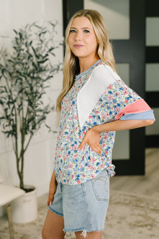 A woman modeling a short-sleeve colorblock top with a blue, pink, and white floral pattern, a solid white ribbed yoke, and pink and blue striped sleeve cuffs, paired with denim shorts in an indoor setting.
