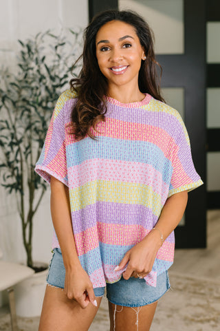 A woman wearing a pastel rainbow striped short-sleeve knit top and denim shorts, standing in a brightly lit indoor setting.