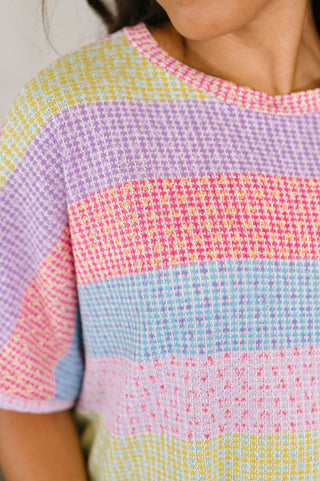 A close-up of a person wearing a short-sleeve knit top with horizontal pastel stripes in pink, blue, purple, and yellow.