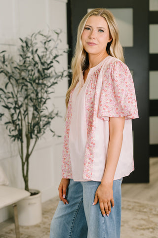 A woman modeling a light pink blouse with a vertical stripe pattern on the front and a pink floral print on the short sleeves and side panels, paired with light blue jeans in an indoor setting.