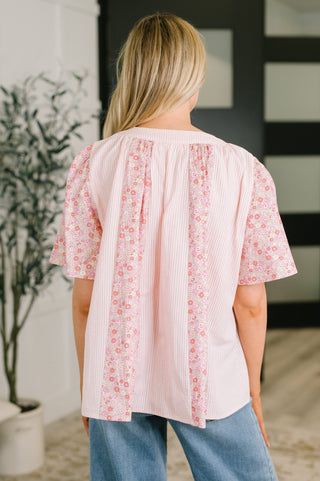 A woman shown from the back wearing a light pink blouse with vertical floral panels and pleated detailing, paired with blue jeans in an indoor setting.