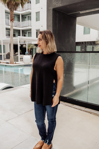 Woman in black sleeveless top and blue jeans standing by a poolside.
