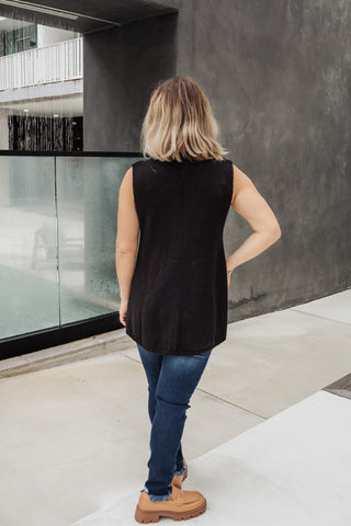 Person wearing a black sleeveless top and blue jeans standing in front of a modern building.
