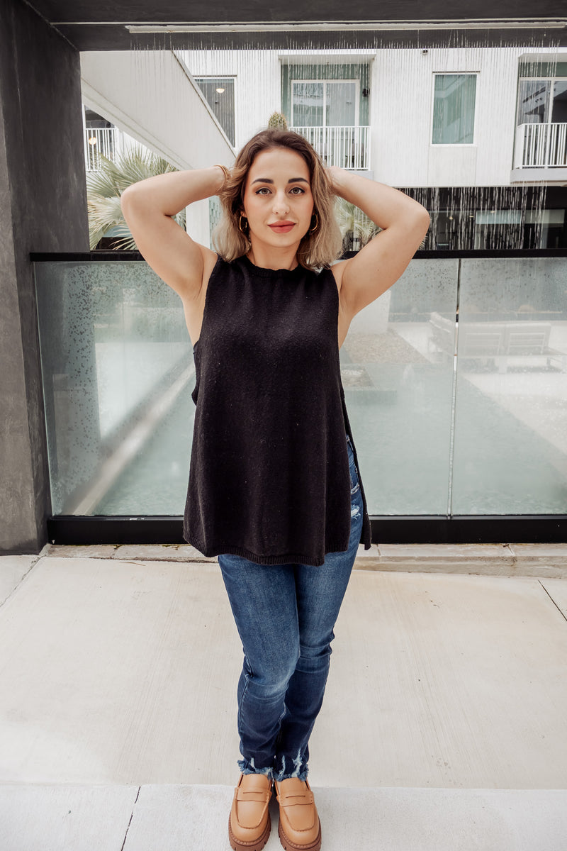Woman wearing a black sleeveless top and blue jeans standing in front of a modern building.
