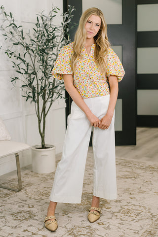 Woman in a floral blouse and white pants standing in a room with a plant and bench.