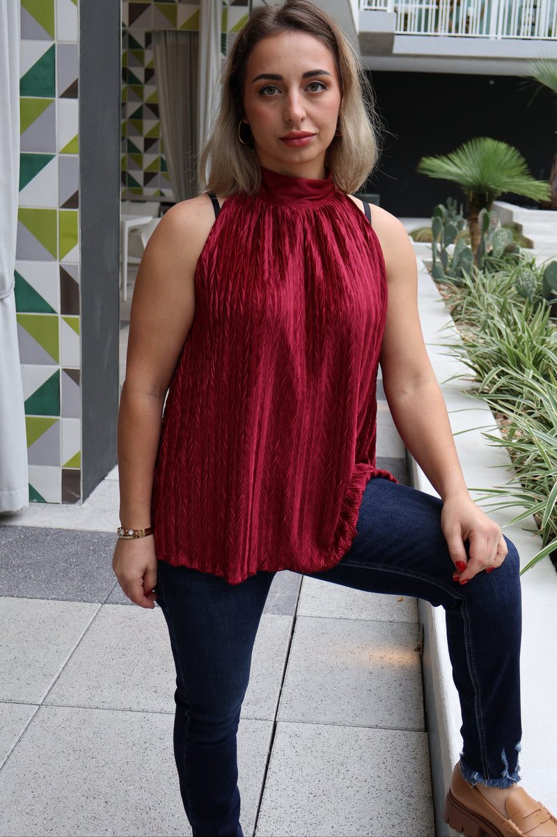 Woman in a red top and blue jeans standing in an indoor setting with plants and a geometric-patterned wall.