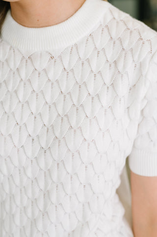 Close-up of a person wearing a white short-sleeve knit top with an openwork scalloped or fish-scale pattern across the chest and shoulders.