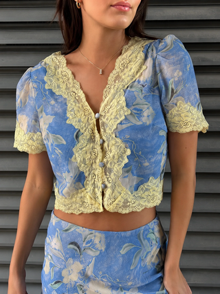 A woman wearing a light blue floral sheer blouse with pale yellow lace trim and puff sleeves, paired with a matching floral skirt, set against a gray slatted wall.